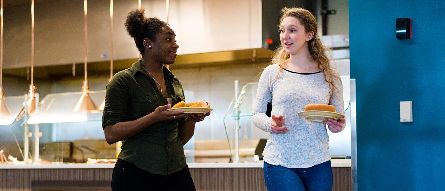 two female students carrying plates of food in a dining hall