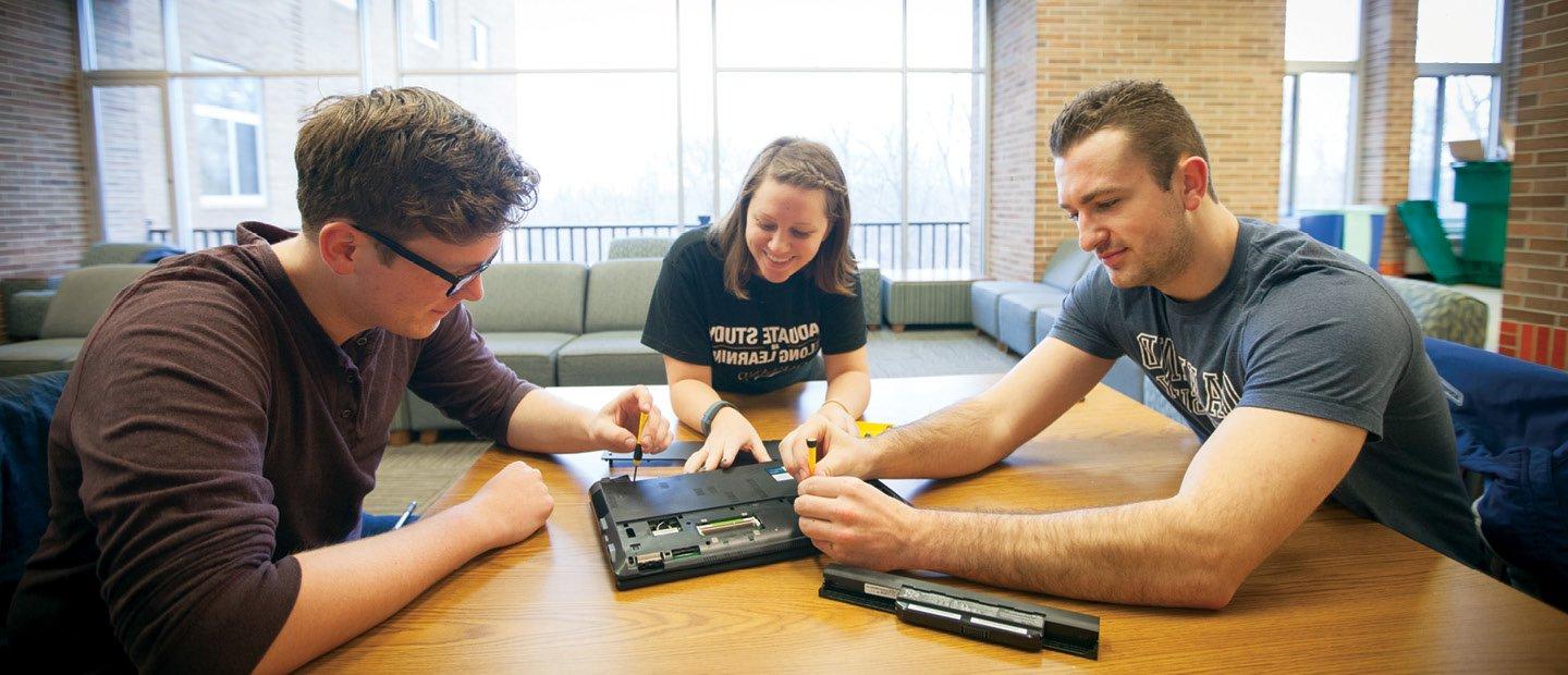 三个学生围坐在一张桌子旁，用螺丝刀检查一台笔记本电脑